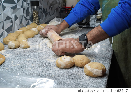 Making unleavened flatbreads. A man's hands roll out roti dough with a rolling pin. The surface is dusted with multigrain flour. Several balls of dough lie nearby. A gas stove is in the background. Making unleavened flatbreads. A man's hands roll out roti dough with a rolling pin. The surface is dusted with multigrain flour. Several balls of dough lie nearby. A gas stove is in the background. 134462347