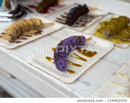 Banana coated with crumbs and chocolate sauce on paper plate. Street dessert detail, sweet snack presentation, and festive food mood 134462939
