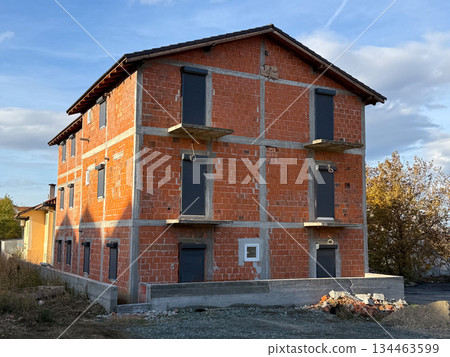 Unfinished brick residential building with closed shutters and raw facade under open sky. Real estate development, housing investment, construction progress, urban growth, and future property 134463599