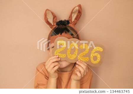 A child with horse ears hides his face on the golden numbers of the 2026 new year. Congratulations on the holiday A child with horse ears hides his face on the golden numbers of the 2026 new year. Congratulations on the holiday 134463745