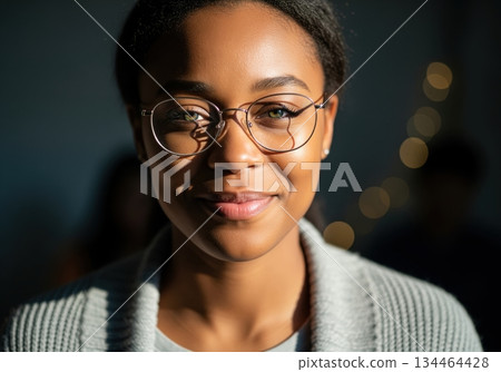 Close up portrait of a smiling young black woman wearing round glasses and a grey cardigan 134464428