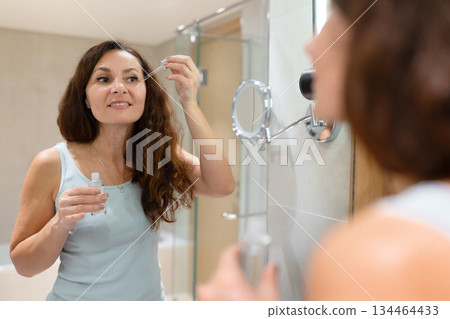 Adult caucasian woman applying cosmetic serum under eyes near bathroom mirror 134464433