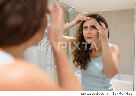 Adult woman checking forehead lines with fingers while looking in bathroom mirror 134464452