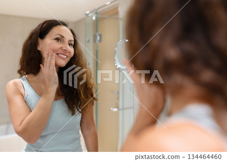 Smiling adult woman touching her cheek while looking in bathroom mirror Smiling adult woman touching her cheek while looking in bathroom mirror 134464460