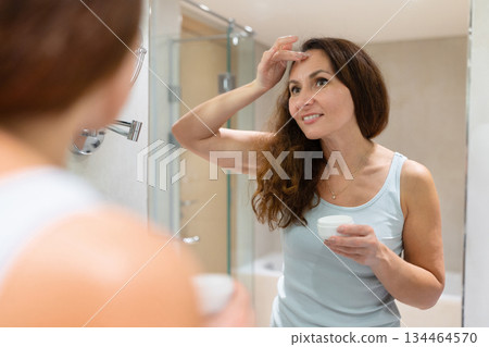 Adult woman applying face cream on forehead and smiling at mirror reflection 134464570