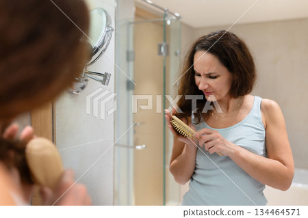 Adult woman brushing dry split hair ends while standing near bathroom mirror Adult woman brushing dry split hair ends while standing near bathroom mirror 134464571
