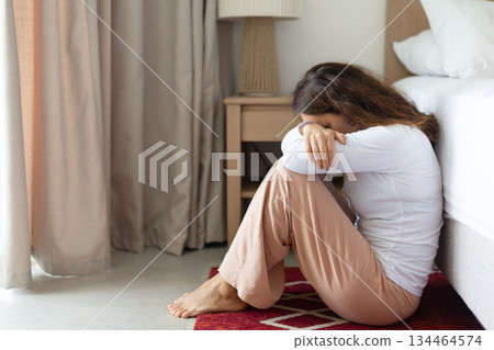 Upset woman sitting on floor near bed resting head on arms 134464574