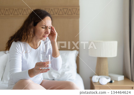 Woman holding glass of water and touching temple, experiencing headache on bed Woman holding glass of water and touching temple, experiencing headache on bed 134464624