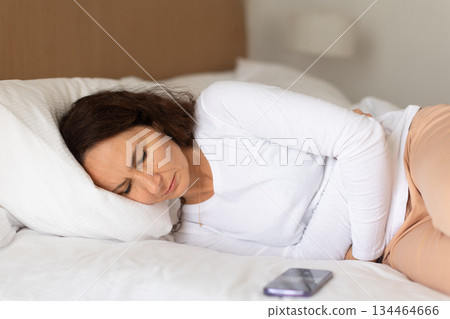 Adult woman lying on side in bed holding stomach, experiencing abdominal pain at home 134464666