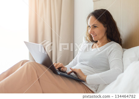 Smiling adult woman working on laptop while reclining on bed 134464782