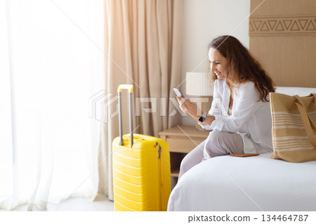 Woman sitting on bed, focused on smartphone with yellow suitcase beside her Woman sitting on bed, focused on smartphone with yellow suitcase beside her 134464787