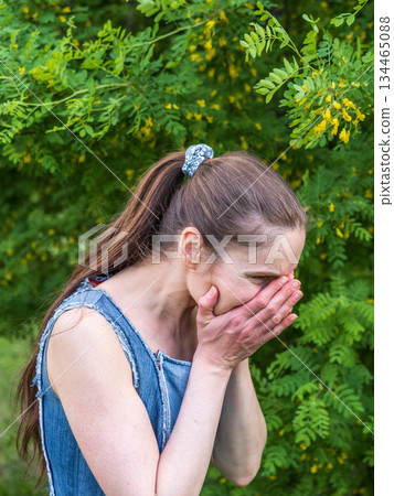 Young woman sneezes in the park against the background of a flowering tree. Allergy to pollen concept. Young woman sneezes in the park against the background of a flowering tree. Allergy to pollen concept. 134465088