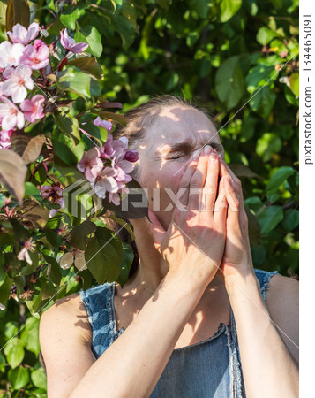 Young woman sneezes in the park against the background of a flowering tree. Allergy to pollen concept. Young woman sneezes in the park against the background of a flowering tree. Allergy to pollen concept. 134465091