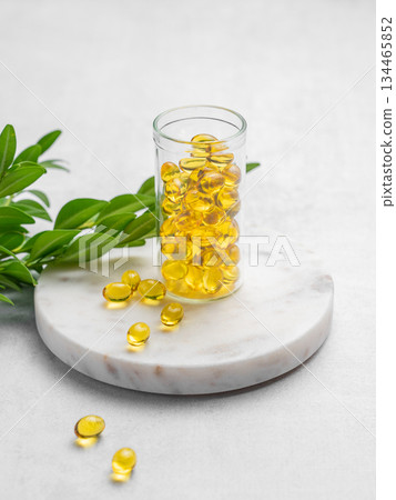 Yellow softgel vitamins capsules in a bottle on a marble standon a light background with green leaf. Yellow softgel vitamins capsules in a bottle on a marble standon a light background with green leaf. 134465852