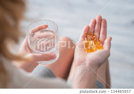 A close-up of a person's hands holding yellow softgels in one palm and a glass of water in the other A close-up of a person's hands holding yellow softgels in one palm and a glass of water in the other 134465857