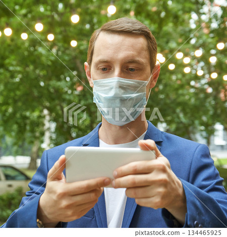 Caucasian male adult wearing face mask using tablet outdoors with greenery 134465925
