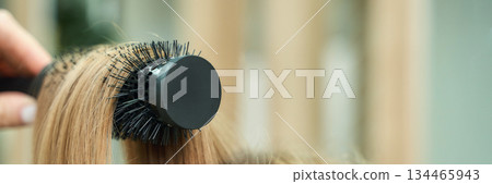 Close-up of round hairbrush styling blonde hair with hand indoors Close-up of round hairbrush styling blonde hair with hand indoors 134465943