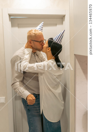 Couple Kissing Birthday Party Doorway: Romantic indoor celebration; man and woman wearing party hats embrace and kiss near a door during a birthday. Couple Kissing Birthday Party Doorway: Romantic indoor celebration; man and woman wearing party hats embrace and kiss near a door during a birthday. 134465970