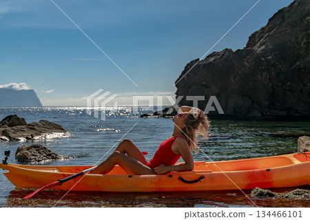 Woman kayak beach, relaxing on an orange kayak near a rocky coast on a sunny summer day 134466101