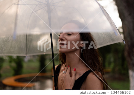 Woman, rain, umbrella. Serene woman enjoying a peaceful moment under a clear umbrella outdoors. 134466102