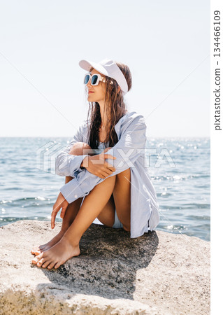 Woman Ocean Relaxing: Barefoot woman sits on coastal rock, enjoying sunny summer day by blue sea. Woman Ocean Relaxing: Barefoot woman sits on coastal rock, enjoying sunny summer day by blue sea. 134466109