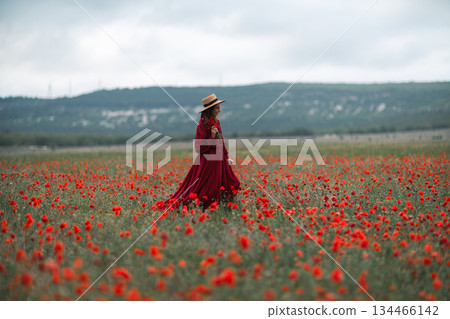 Woman poppy field walking in elegant red dress through summer meadow landscape 134466142