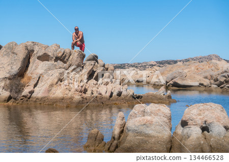 Person standing on rocky seaside near calm sea water. Travel lifestyle, exploration and connection with nature during a summer vacation, Sardinian, La Maddalena, Italy 134466528