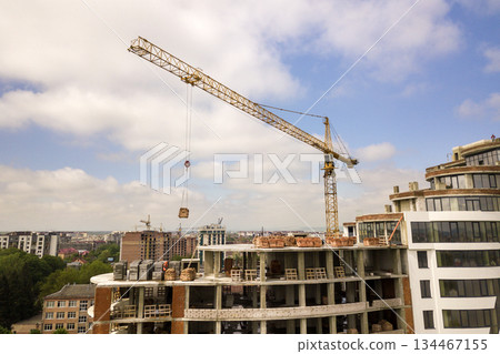 Apartment or office tall building under construction. Brick walls, glass windows, scaffolding and concrete support pillars. Tower crane on bright blue sky copy space background. Apartment or office tall building under construction. Brick walls, glass windows, scaffolding and concrete support pillars. Tower crane on bright blue sky copy space background. 134467155
