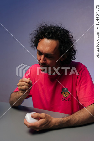 Vertical portrait of handsome young adult male with curly hair trimming fingernails using nail clipper, sitting at gray table with purple backdrop at home. Concept of healthy lifestyle. Vertical portrait of handsome young adult male with curly hair trimming fingernails using nail clipper, sitting at gray table with purple backdrop at home. Concept of healthy lifestyle. 134467579
