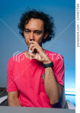 Vertical portrait of man with curly hair wearing pink t-shirt using nasal spray to alleviate allergy symptoms or congestion, sitting at a desk on isolated background. Concept of healthy lifestyle. Vertical portrait of man with curly hair wearing pink t-shirt using nasal spray to alleviate allergy symptoms or congestion, sitting at a desk on isolated background. Concept of healthy lifestyle. 134467580