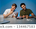 Portrait of laughing two young men sitting at table with drugs, white powder, pills, cake, money and blue light in background. Concept of party atmosphere with substance abuse. 134467583