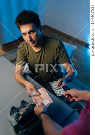 Serious man exchanging Russian rubles for dollars using money counting machine on table at night, looking at camera, illuminated by soft blue light, capturing essence of financial transactions. 134467585