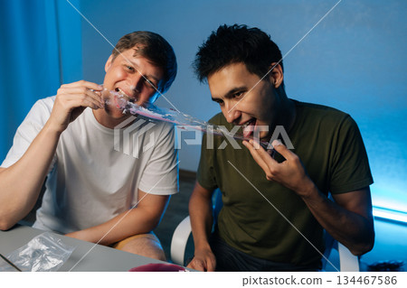 Two young men consuming cake likely containing psychedelic drugs during party at home, showcasing risky and potentially dangerous behavior. Concept of party atmosphere with substance abuse. 134467586