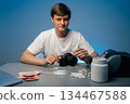 Cocaine dealer sitting at table, displaying drug paraphernalia, cash and pills, holding rolled banknote, looking at camera, studio shot portrait on isolated background. Concept of substance abuse. 134467588