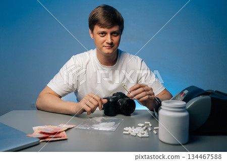 Cocaine dealer sitting at table, displaying drug paraphernalia, cash and pills, holding rolled banknote, looking at camera, studio shot portrait on isolated background. Concept of substance abuse. Cocaine dealer sitting at table, displaying drug paraphernalia, cash and pills, holding rolled banknote, looking at camera, studio shot portrait on isolated background. Concept of substance abuse. 134467588