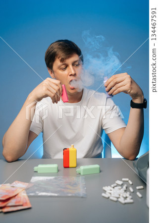 Vertical portrait of drug dealer sitting at desk with scattered cash, pharmaceuticals, smoking electronic heated tobacco device, exhaling vapor against blue background. 134467591