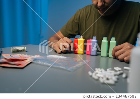 Cropped shot of unrecognizable drug dealer arranging colorful vape pods, pills and cash on desk at night, preparing illegal merchandise for sale with bill counter machine in background. Cropped shot of unrecognizable drug dealer arranging colorful vape pods, pills and cash on desk at night, preparing illegal merchandise for sale with bill counter machine in background. 134467592