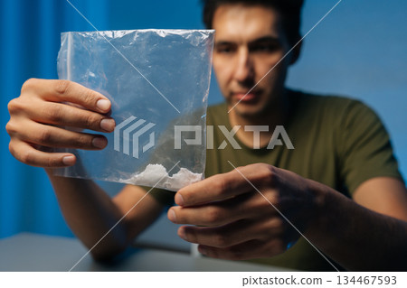 Close-up of of serious drug dealer male preparing dose of cocaine or methamphetamine from plastic bag sitting at table in dark room. Concept of drug dealing or addiction. 134467593
