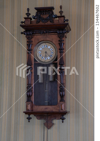 Antique wooden wall clock with carved details, pendulum and weights mounted on striped wallpaper, warm brown tones and vintage craftsmanship Antique wooden wall clock with carved details, pendulum and weights mounted on striped wallpaper, warm brown tones and vintage craftsmanship 134467602