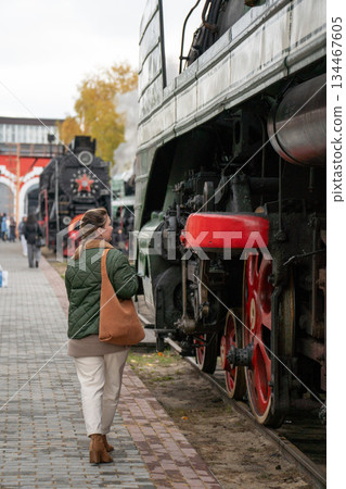 Woman in a green jacket stands beside a vintage steam locomotive with red wheels on an outdoor railway platform, industrial details and autumn mood 134467605