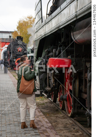 Woman in a green jacket stands beside a vintage steam locomotive with red wheels on an outdoor railway platform, industrial details and autumn mood 134467606