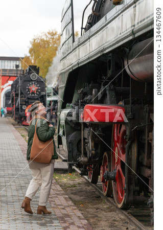Woman in a green jacket stands beside a vintage steam locomotive with red wheels on an outdoor railway platform, industrial details and autumn mood Woman in a green jacket stands beside a vintage steam locomotive with red wheels on an outdoor railway platform, industrial details and autumn mood 134467609