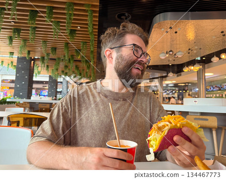 A man with glasses enjoys a taco and drink while sitting inside a food court restaurant. Casual dining, consumer lifestyle, fast food culture and modern gastronomy in everyday leisure. A man with glasses enjoys a taco and drink while sitting inside a food court restaurant. Casual dining, consumer lifestyle, fast food culture and modern gastronomy in everyday leisure. 134467773