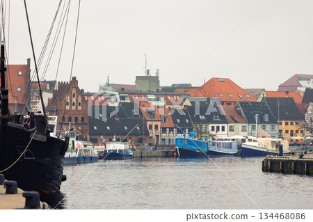 Wismar harbor waterfront moored boats brick building gabled houses red rooftops reflect calm water overcast sky. Hansatic seaport scenery show historic architecture maritime atmosphere urban mood 134468086