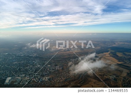 Aerial view from high altitude of distant city covered with puffy cumulus clouds flying by before rainstorm. Airplane point of view of landscape in cloudy weather 134469535