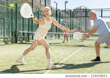Old woman looking back after the ball while playing Padel Tennis in tennis court Old woman looking back after the ball while playing Padel Tennis in tennis court 134470651