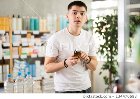 Young male buyer choosing medicinal cough liquid syrup standing between shelves at pharmacy Young male buyer choosing medicinal cough liquid syrup standing between shelves at pharmacy 134470688