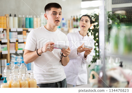 Guy chooses pills in modern pharmacy. Female pharmacist helps with choosing medicine 134470823