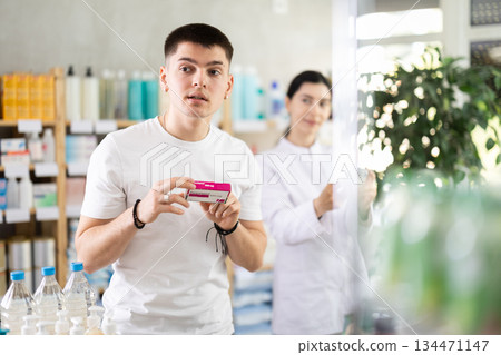 Guy chooses pills paracetamol in modern pharmacy. Female pharmacist helps with choosing medicine 134471147