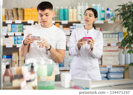 Interested young guy choosing between two boxes of paracetamol antipyretic pills and professional pharmacist woman helping and offering supplements at drugstore 134471297